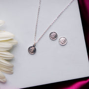 A silver pendant set featuring a pressed coin design, displayed on a white surface with a pair of earrings and some floral elements in the background.